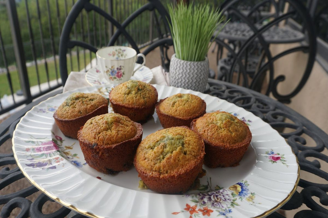 Six golden GoodShoog™ Blueberry-Oat Muffins on a floral china plate on a balcony table with a tea cup in the background